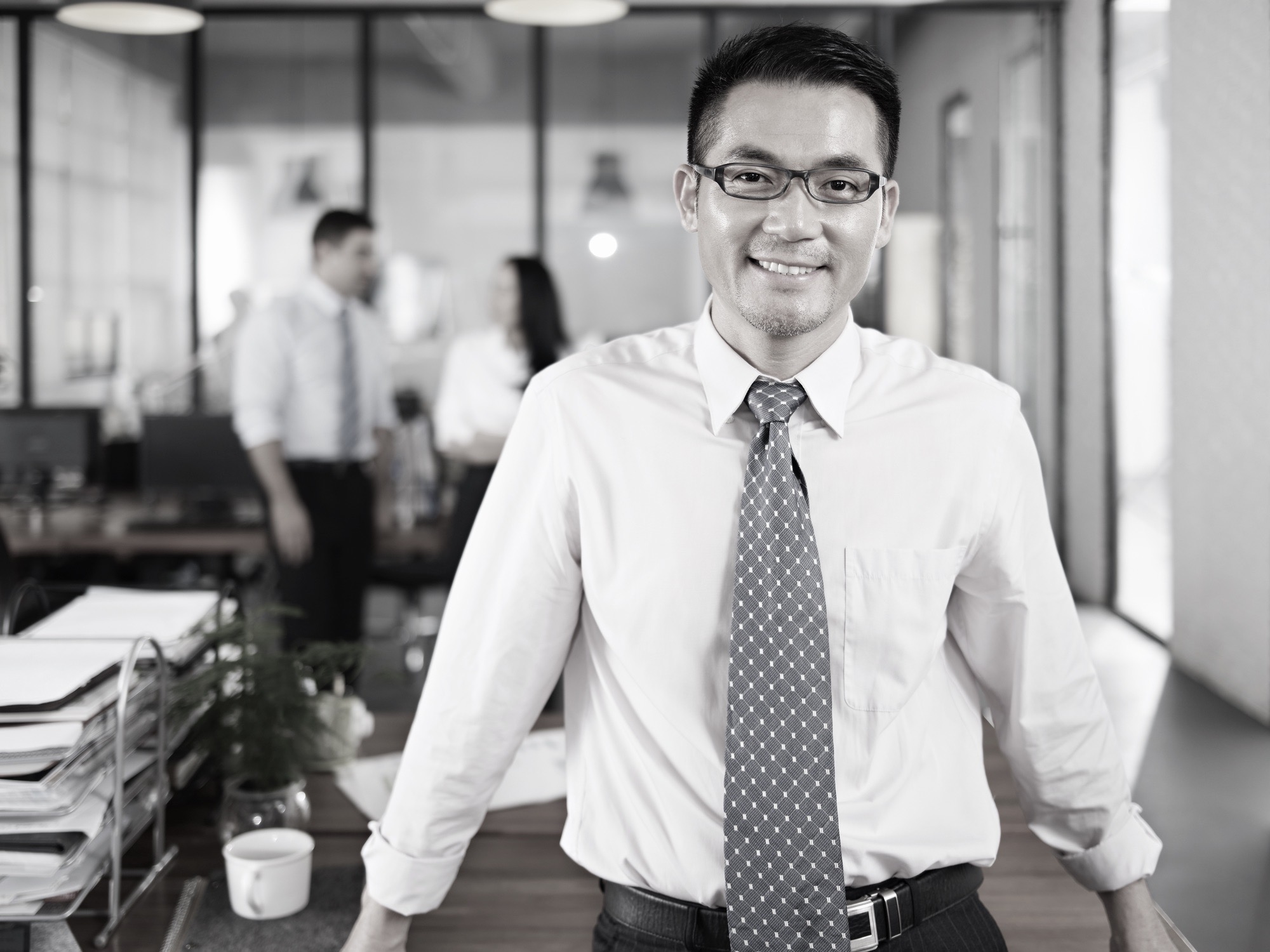 asian businessman standing in office arms crossed with multinational colleagues talking in background.